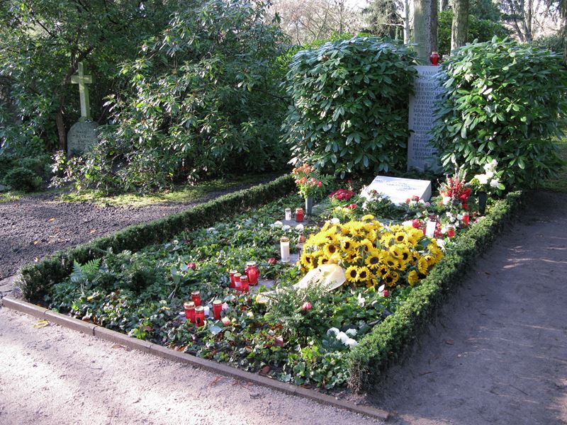 Ein reichlich bepflanztes und gepflegtes Grab, neben dem Grabstein zwei üppige Büsche, auf dem Grab gelb blühende Blumen.