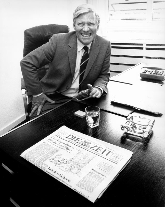 Helmut Schmidt breit lächelnd am Schreibtisch, auf dem eine Ausgabe der Wochenzeitung DIE ZEIT liegt.